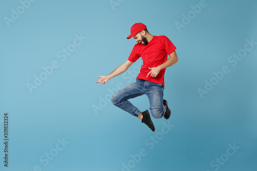 Delivery man in red uniform workwear isolated on blue wall background, studio portrait. Professional male employee in cap t-shirt print working as courier dealer. Service concept. Mock up copy space.