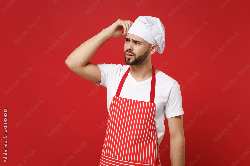 Preoccupied male chef cook or baker man in striped apron white t-shirt toque chefs hat posing isolated on red background. Cooking food concept. Mock up copy space. Looking aside, putting hand on head. Preoccupied male chef cook or baker man in striped apron white t-shirt toque chefs hat posing isolated on red background. Cooking food concept. Mock up copy space. Looking aside, putting hand on head.
