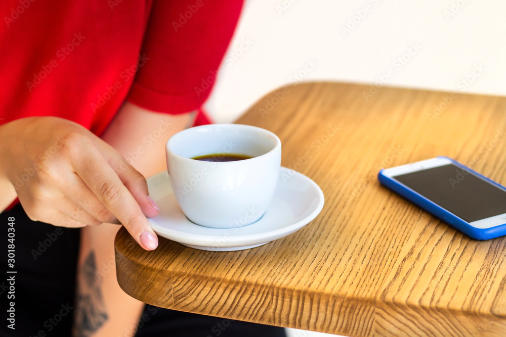 Image of woman's hand holding white ceramic cup with filter coffee, sitting on the wooden table in cafe.