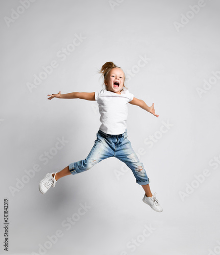 Happy screaming kid girl in blue jeans and white t-shirt is jumping with her arms up spread having fun on white