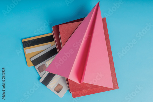Red passports with credit cards lie on a blue background under a pink paper airplane © Pavel Iarunichev
