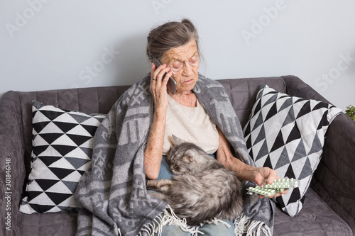 Sick senior woman taking medicine at home. Senior woman talking on mobile phone while looking at medicine in bedroom. pensioner studying medicine calling doctor while sitting on sofa with cat photo