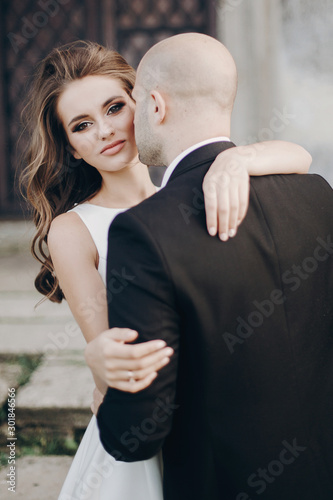 Gorgeous bride and groom embracing in evening sunlight near old castle in park. Romantic moment. Stylish wedding couple gently hugging and smiling