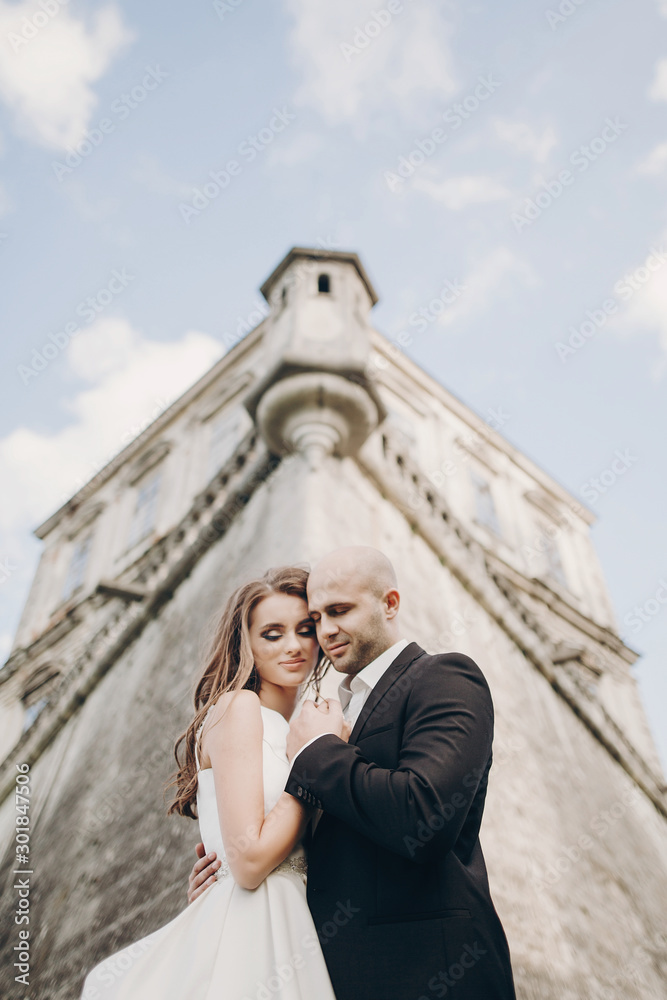 Gorgeous wedding couple embracing in sunlight near old castle in beautiful park. Stylish beautiful bride and groom gently hugging on background of ancient building and nature