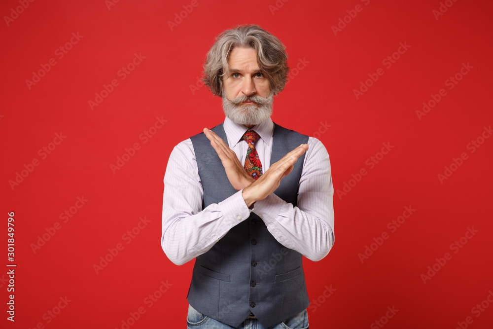 Serious elderly gray-haired mustache bearded man in classic shirt vest colorful tie isolated on red background. People lifestyle concept. Mock up copy space. Showing stop gesture with crossed hands.
