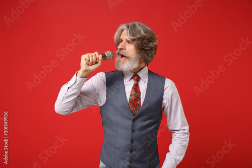 Elderly gray-haired mustache bearded man in classic shirt vest and colorful tie isolated on red wall background, studio portrait. People lifestyle concept. Mock up copy space. Sing song in microphone. photo