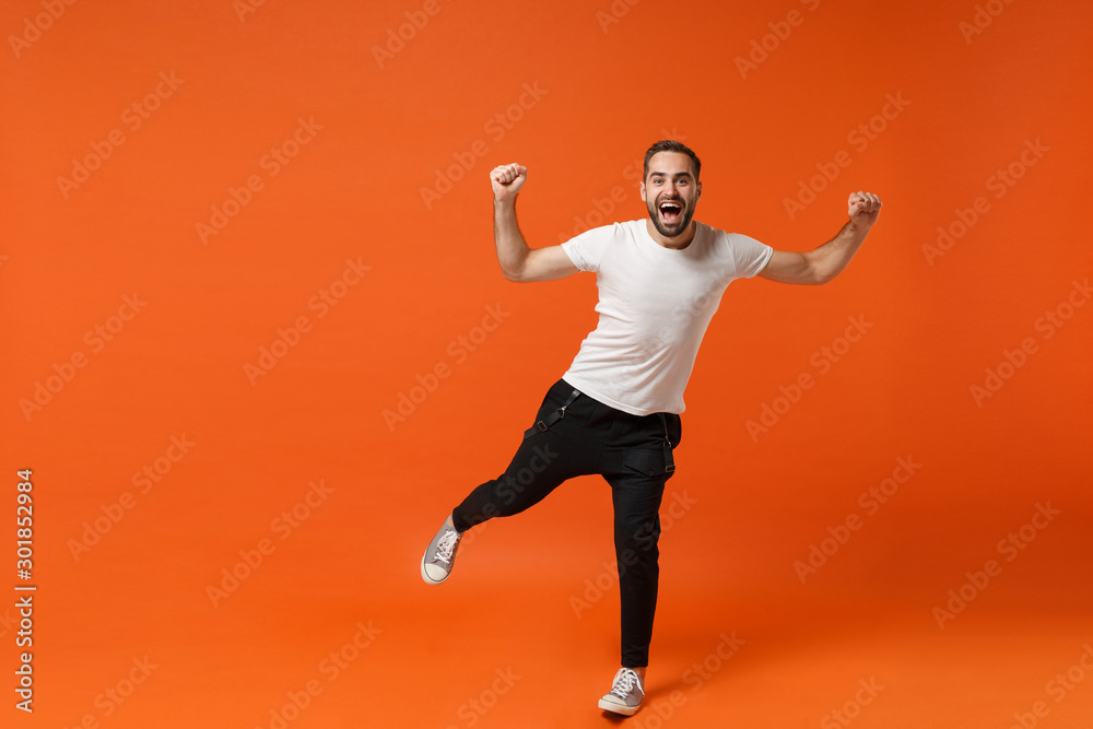 Cheerful young man in casual white t-shirt posing isolated on bright orange wall background studio portrait. People sincere emotions lifestyle concept. Mock up copy space. Clenching fists like winner.