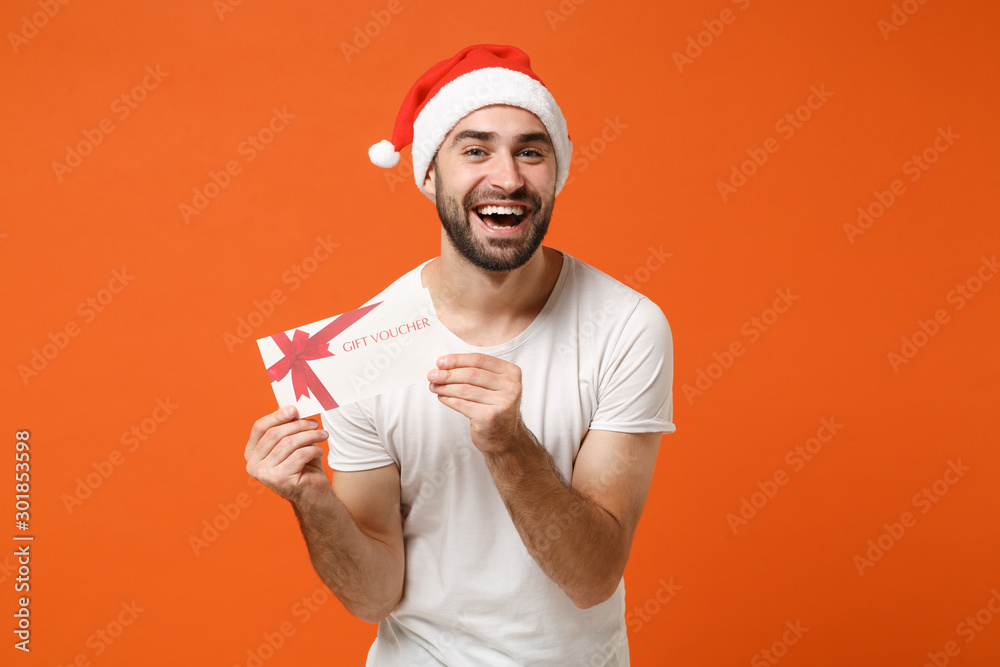 Laughing young Santa man in white t-shirt, Christmas hat posing isolated on orange background in studio. Happy New Year 2020 celebration holiday concept. Mock up copy space. Holding gift certificate. Laughing young Santa man in white t-shirt, Christmas hat posing isolated on orange background in studio. Happy New Year 2020 celebration holiday concept. Mock up copy space. Holding gift certificate.