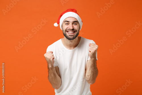 Smiling young Santa man in white t-shirt, Christmas hat posing isolated on orange background studio portrait. Happy New Year 2020 celebration holiday concept. Mock up copy space. Doing winner gesture.