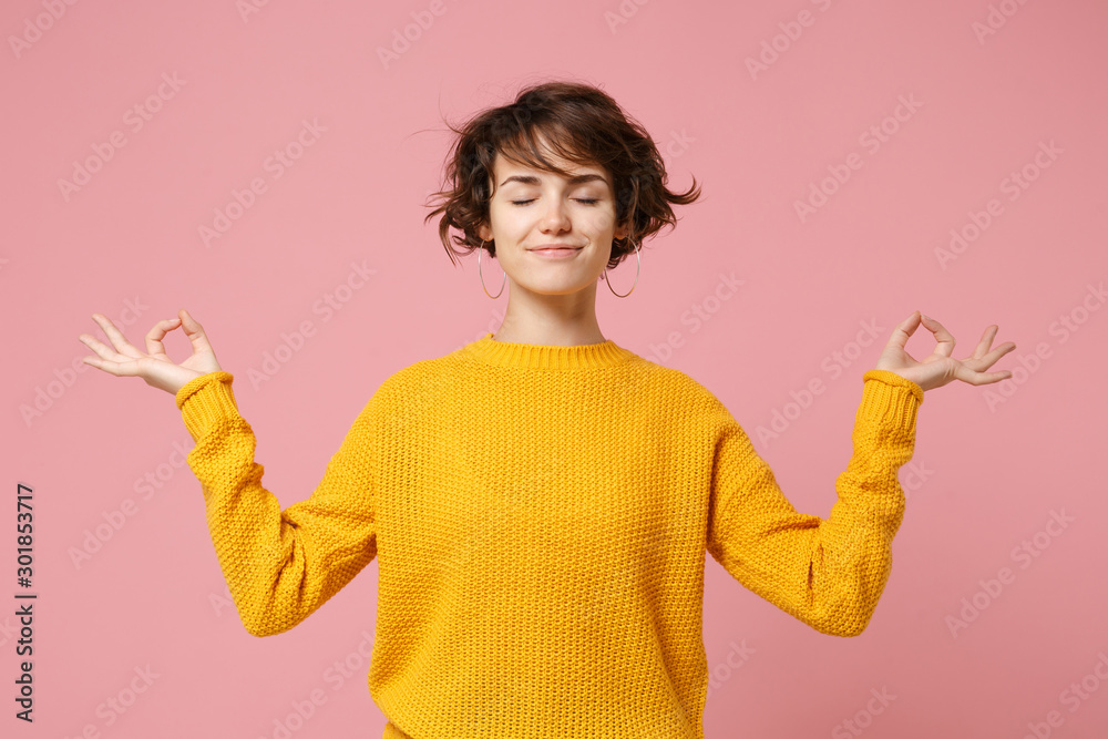 Young brunette woman girl in yellow sweater posing isolated on pastel pink background. People lifestyle concept. Mock up copy space. Hold hands in yoga gesture relaxing meditating keeping eyes closed. - obrazy, fototapety, plakaty 