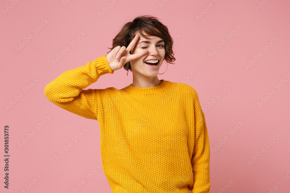 Cheerful young brunette woman girl in yellow sweater posing isolated on pastel pink wall background, studio portrait. People lifestyle concept. Mock up copy space. Blinking, showing victory sign.