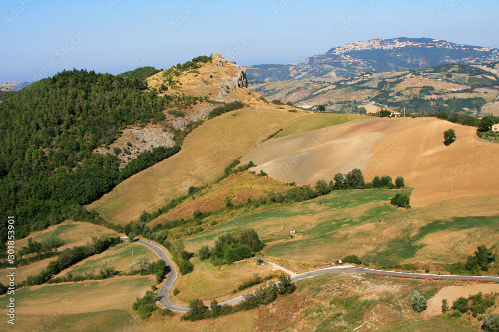 Beautiful Italian landscape, gray road ribbon on a green-brown background of the foothills. Sunny summer day, blue cloudless sky, mountains in the air haze.