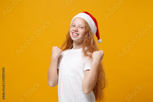 Happy little ginger kid Santa girl 12-13 years old in white t-shirt, Christmas hat isolated on yellow background. New Year 2020 celebration holiday concept. Mock up copy space. Doing winner gesture. © ViDi Studio