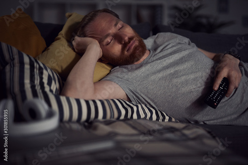 Portrait of bearded adult man sleeping on sofa while watching TV at night, copy space