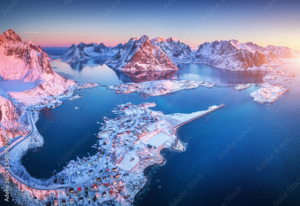 Aerial view of Reine at sunrise in winter. Top view of Lofoten islands, Norway. Landscape with blue sea, snowy mountains, high rocks, village with buildings, rorbu, blue sky, reflection in water - obrazy, fototapety, plakaty 