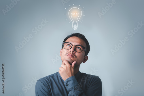 Thinking asian man in glasses looking up with light idea bulb above head on gray background.creative idea.Concept of idea and innovation