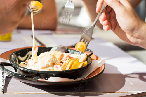 Hands holding cutlery - forks, picking hot fried potatoes with cheese and sauce in an iron pan on a table in the sunlight from a restaurant window, close-up,View from above.The process of taking food 