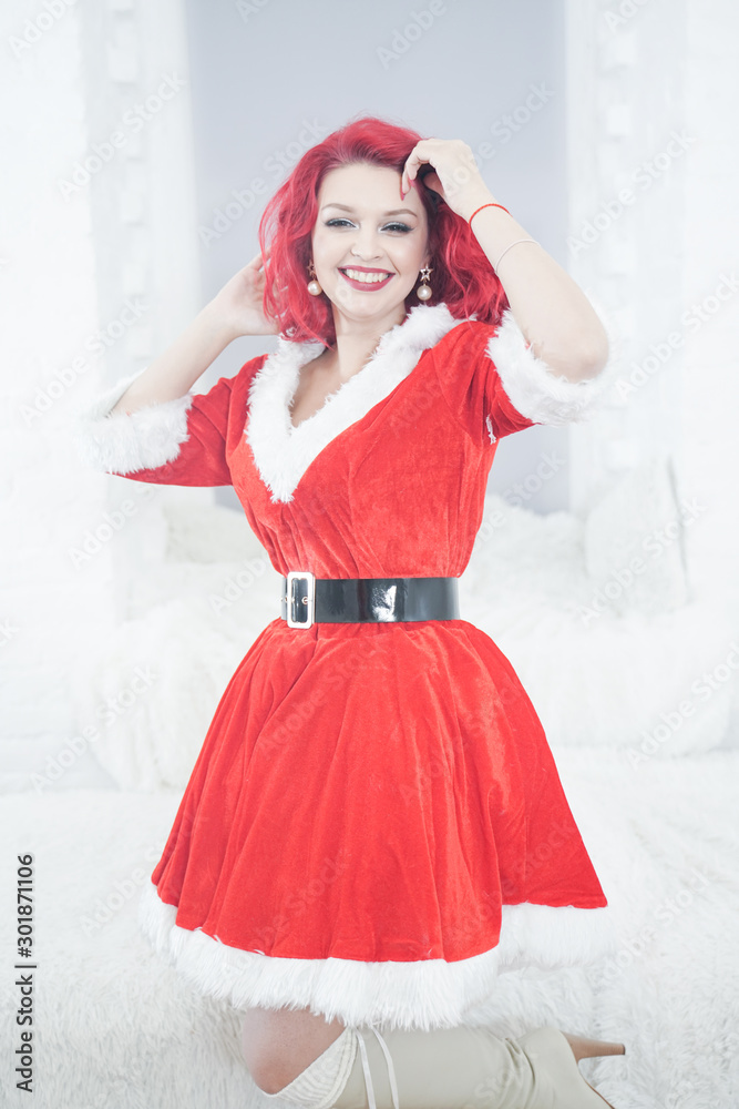 pretty redhaired young stylish woman in velvet santa dress with fur posing on white room background near the cozy window pretty redhaired young stylish woman in velvet santa dress with fur posing on white room background near the cozy window