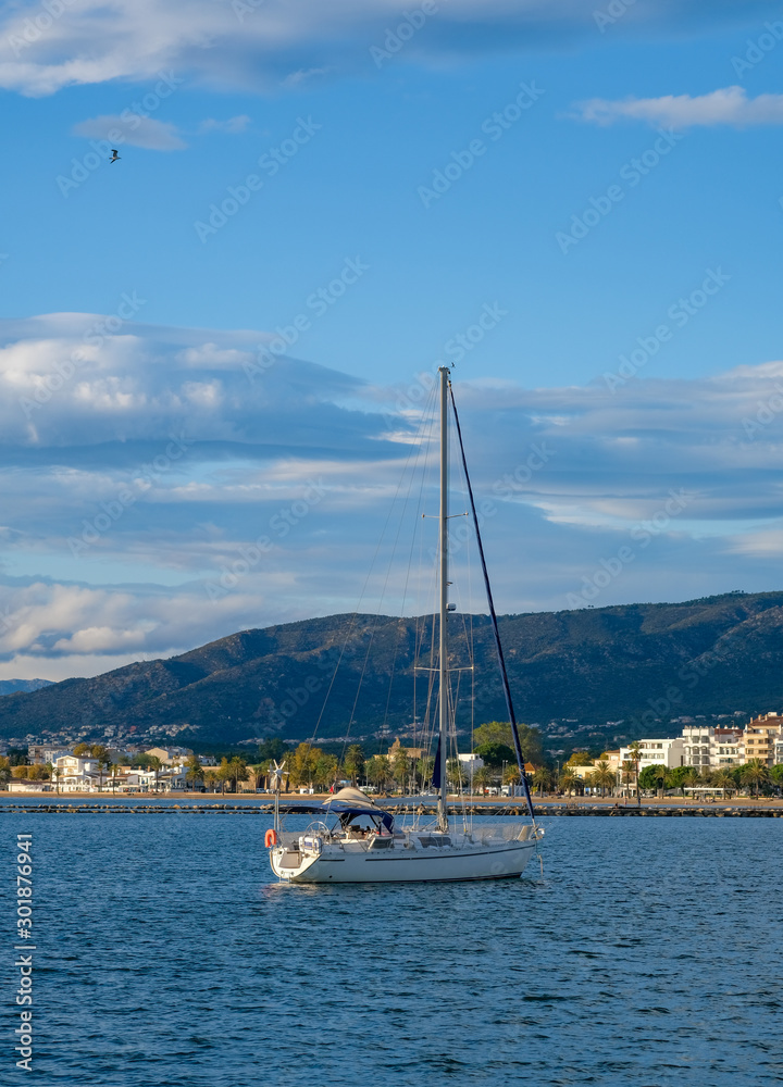 Sailing yacht, sailboat in the bay on city landscape and cloudy blue sky background. The boat is anchored in the bay with a calm sea. View from the sea to the coastal city. Roses, Catalonia, Spain.