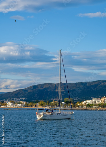 Sailing yacht, sailboat in the bay on city landscape and cloudy blue sky background. The boat is anchored in the bay with a calm sea. View from the sea to the coastal city. Roses, Catalonia, Spain. © Olga