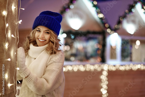 Smiling woman with garlands and holiday lights on festive Christmas or New Year fair. Lady wearing classic stylish winter knitted sweater and mittens © Joe-L