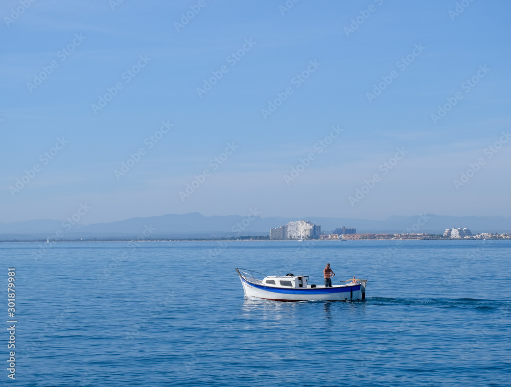 Fisherman on a small boat at sea fishing on coastal city and blue sky background. Fishing in the bay by a city dweller. Calm water in sunny day. Roses, Catalonia, Spain. 