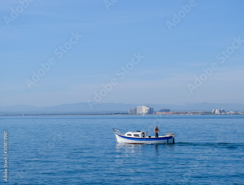 Wallpaper Mural Fisherman on a small boat at sea fishing on coastal city and blue sky background. Fishing in the bay by a city dweller. Calm water in sunny day. Roses, Catalonia, Spain.  Torontodigital.ca
