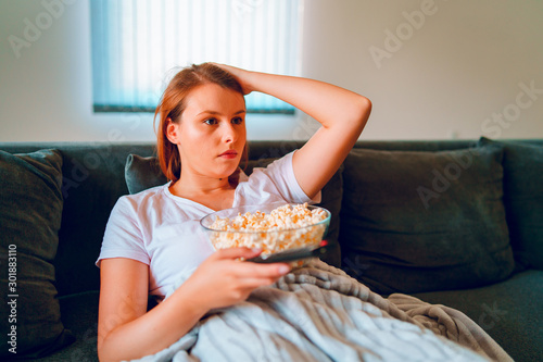Young woman sitting on the bed sofa at home  lying with a popcorn bowl watching TV covered with blanket at her apartment alone enjoying movies or series