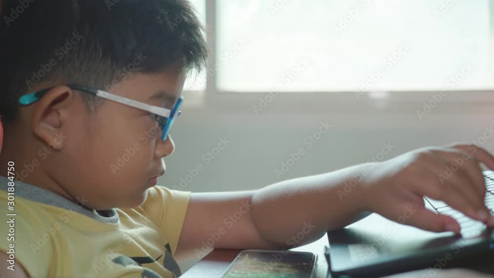 Close up of Happy Asian child boy wear eye glasses play games with laptops computer and watch cartoons on a smartphone  in a room that has sunlight from the window at home.