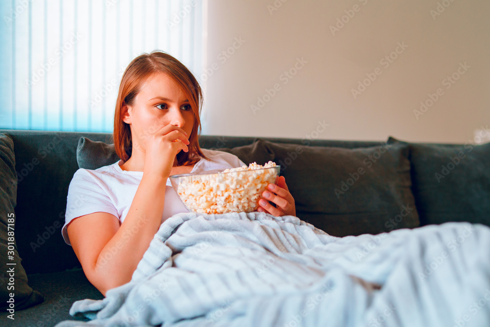 Young woman sitting on the bed sofa at home, lying with a popcorn bowl watching TV covered with blanket at her apartment alone enjoying movies or series Young woman sitting on the bed sofa at home, lying with a popcorn bowl watching TV covered with blanket at her apartment alone enjoying movies or series
