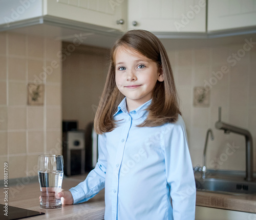 Portrait of a little caucasian baby girl holding a glass of tap clean water. Kitchen faucet. Cute kid pouring fresh water from filter tap. Indoors. Healthy life concept