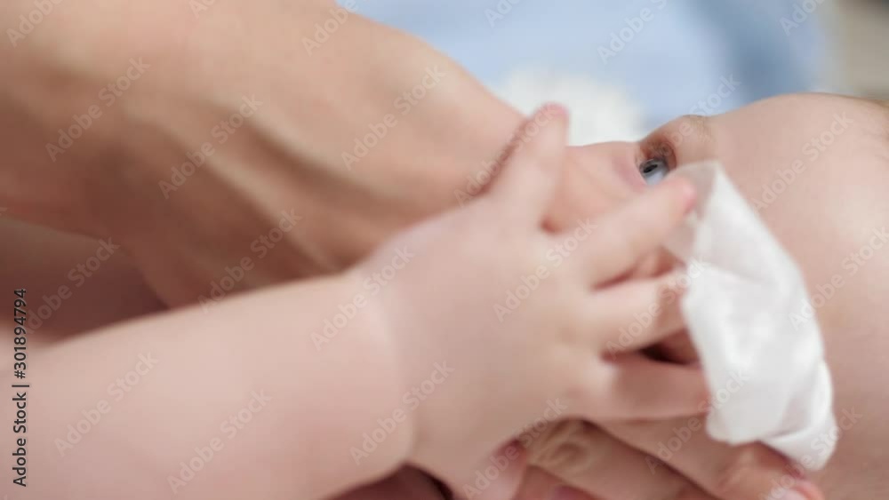 Mom wipes baby's face with wet wipe. Child lies on white blanket on blue background, mother wipes his face with wet cloth. Slow motion Mom wipes baby's face with wet wipe. Child lies on white blanket on blue background, mother wipes his face with wet cloth. Slow motion