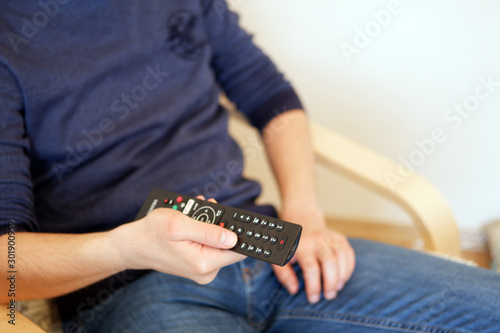 Young man sitting on chair while switching TV channels. Focus on remote control.