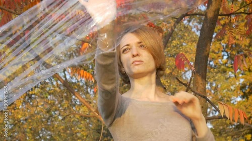 Young woman with short blonde hair is looking through a plastic bag, polyethylene wrap standing outdoors durind sunny autumn day near the tree with yellow and red leaves. photo