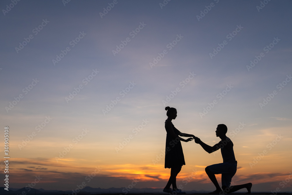 Silhouette couple man down on one knee proposing to woman under sunset sky background