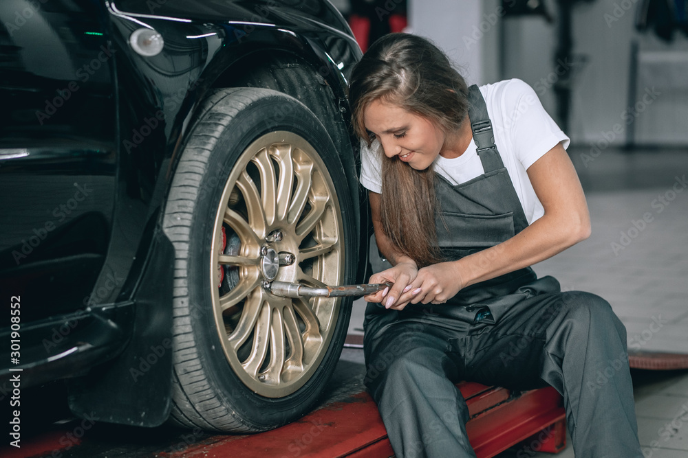 Obraz premium A beautiful girl in a white T-shirt and black jumpsuit is sitting near a black car removing the wheel in the service station