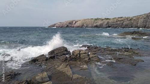 Low angle pan shot rocky bay in Saint Peter island at coastal area named Conca with emerald sea water crashing on the rocks and huge cliff at horizon overlooking the sea, Sardinia region, Italy