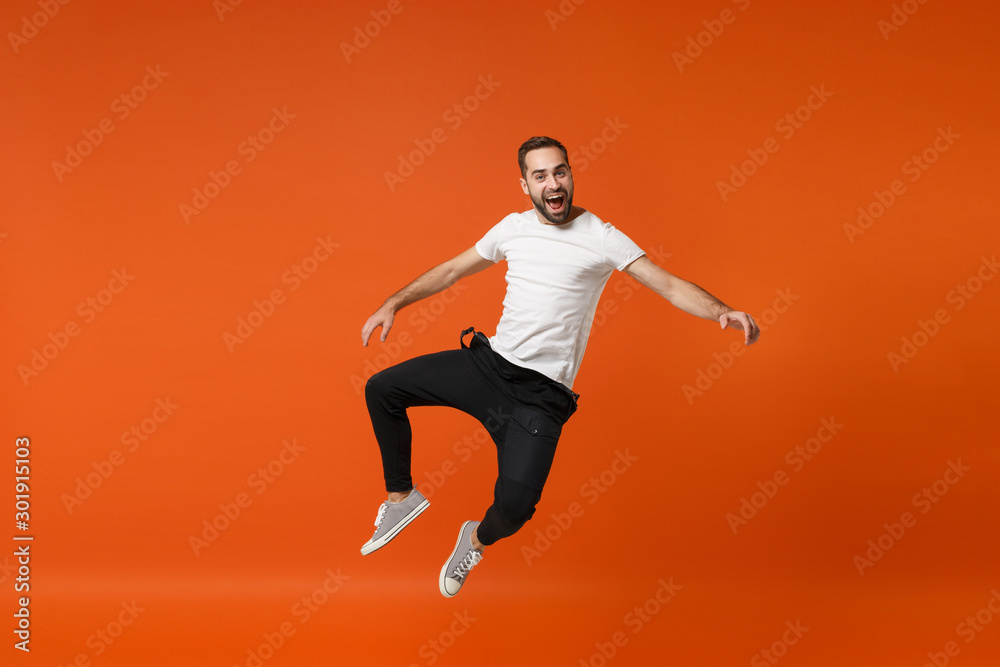Cheerful excited young man in casual white t-shirt posing isolated on bright orange wall background studio portrait. People lifestyle concept. Mock up copy space. Having fun, fooling around, jumping.