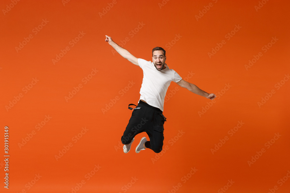 Joyful young man in casual white t-shirt posing isolated on orange wall background studio portrait. People sincere emotions lifestyle concept. Mock up copy space. Having fun jumping, spreading hands.