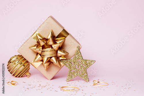Christmas greeting card composition. Craft paper gift with golden ball  confetti star and gold decoration on pink background. Copy space