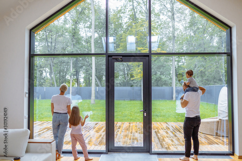 Back of Parents with children look at panoramic window together, relaxing together, spending time at home. Modern house interior