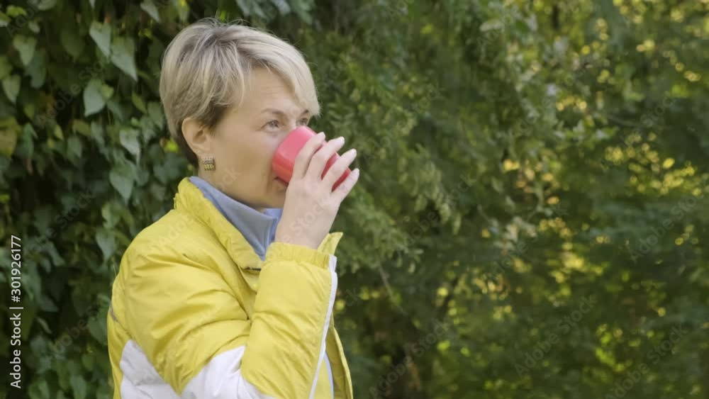 Adult woman drinks hot tea in park on background of green foliage of trees. Adult woman drinks hot tea in park on background of green foliage of trees.
