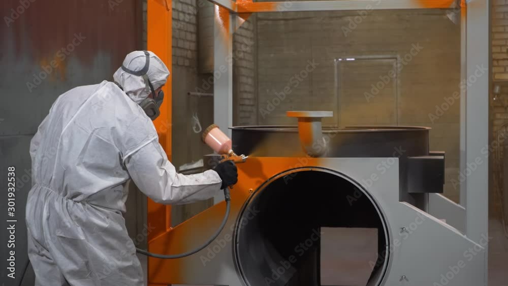 Factory worker paints frame of mobile boiler in orange using spray gun, paint, industrial painting, spacious workshop, man in chemical protection suit, respirator on face, hazardous work, production Factory worker paints frame of mobile boiler in orange using spray gun, paint, industrial painting, spacious workshop, man in chemical protection suit, respirator on face, hazardous work, production