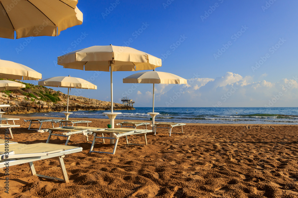 Umbrella beach. The most beautiful beaches of Apulia: Gusmay beach is surrounded by the Mediterranean scrub. In the background a characteristic trabucco (or trebuchet), Italy. Umbrella beach. The most beautiful beaches of Apulia: Gusmay beach is surrounded by the Mediterranean scrub. In the background a characteristic trabucco (or trebuchet), Italy.