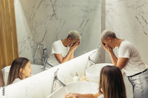 Young woman with her kid in bathroom washing faces. Morning together with child. photo
