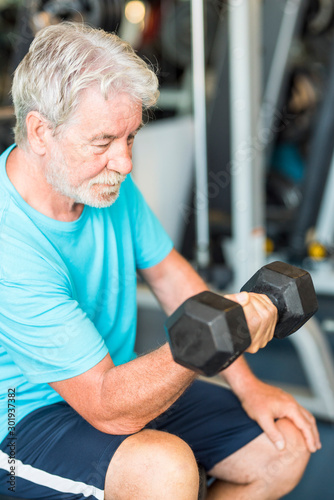 mature man at the gym training his body and his biceps sitting in a bench with a dumbbell in his hand - healthy and fitness senior lifestyle