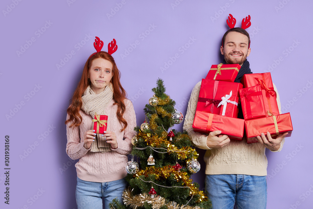 Tall handsome man satisfied by great number of gifts, holding presents with pleasure, thankful to friends, upset girl with small christmas gift in hands expressing disappointment Tall handsome man satisfied by great number of gifts, holding presents with pleasure, thankful to friends, upset girl with small christmas gift in hands expressing disappointment