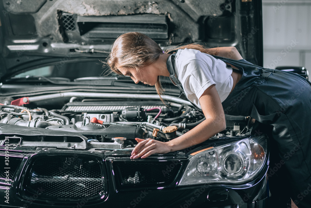 A brunette in a black jumpsuit and a white t-shirt near the open hood. Young female in the garage is smiling at the camera and lowered gaze. car repair concept