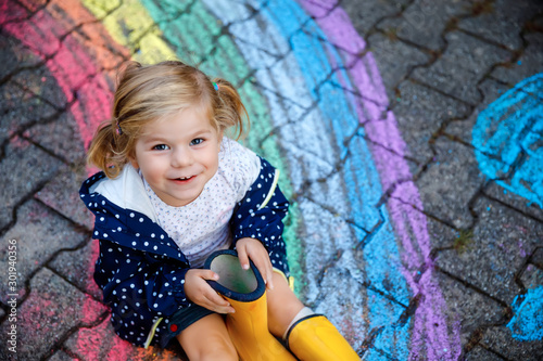 Foto Happy little toddler girl in rubber boots with rainbow sun and clouds with rain painted with colorful chalks on ground or asphalt in summer