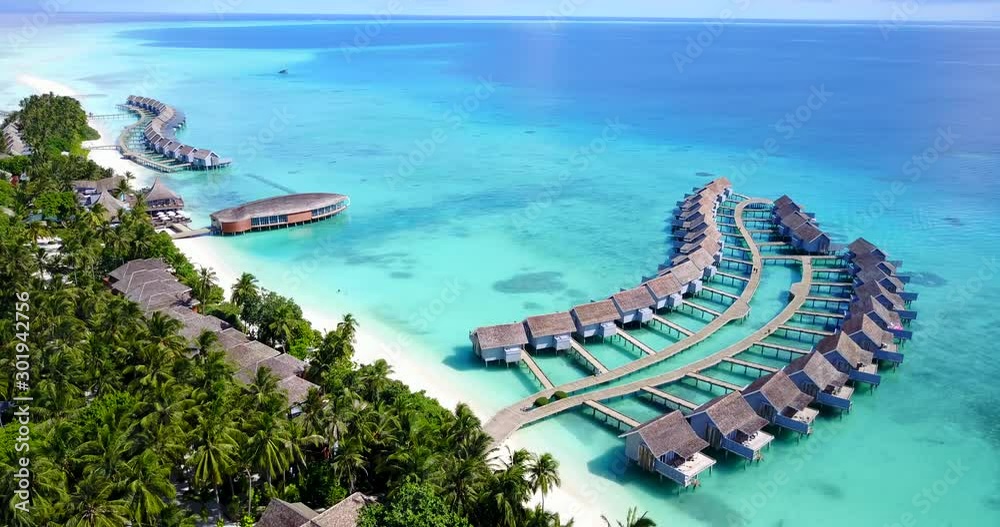 Aerial shot pan of a drone flying above a tropical island with crystal clear waters and an over water bungalow complex in Maldives